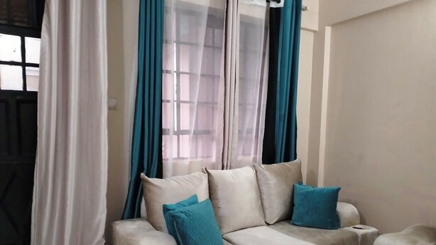 A living room with a beige sofa set, two blue cushions, and a window with teal curtains. - Paradise Apartments Embu (Embu)