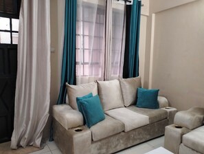 Een woonkamer met een beigekleurige zithoek, twee blauwe kussens en een raam met turquoise gordijnen.