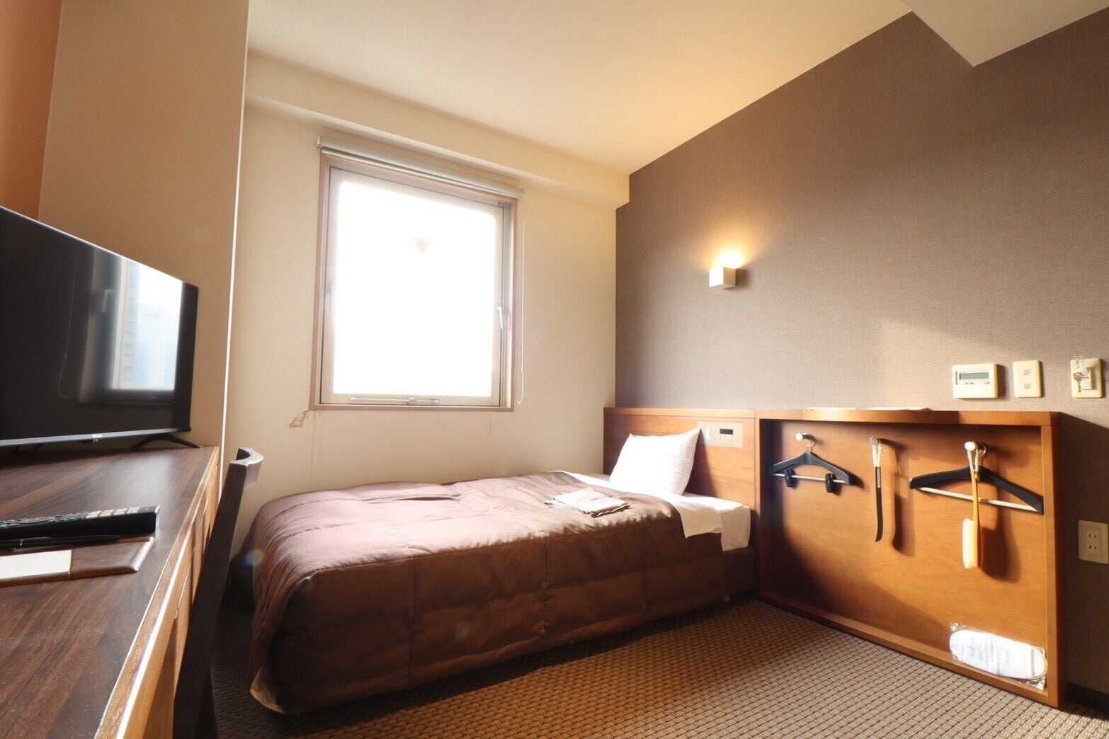 Habitación de hotel con cama, televisor, ventana y perchero en la pared.