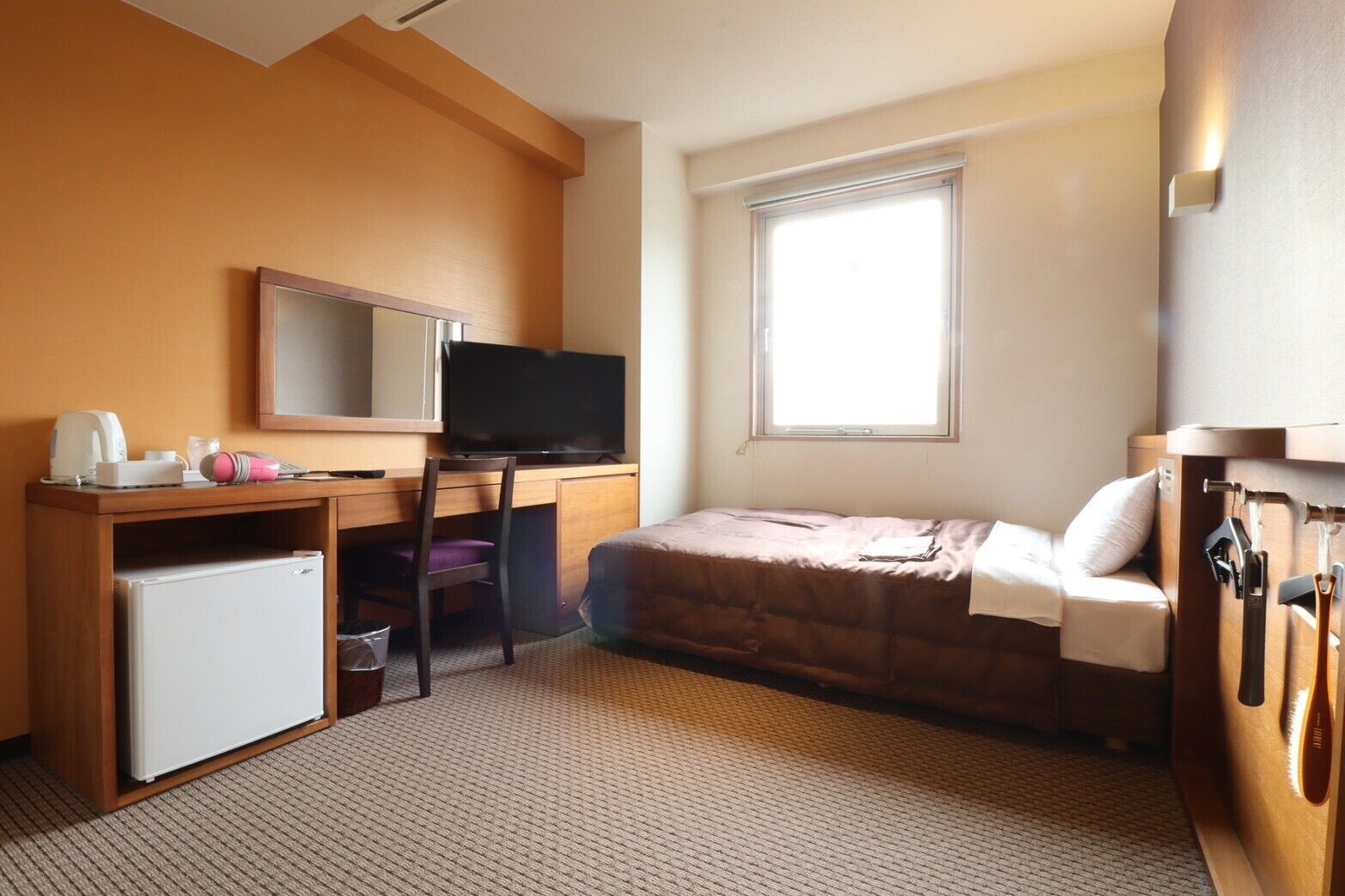 A hotel room with a bed, a desk with a television, a small refrigerator, and a window providing natural light.