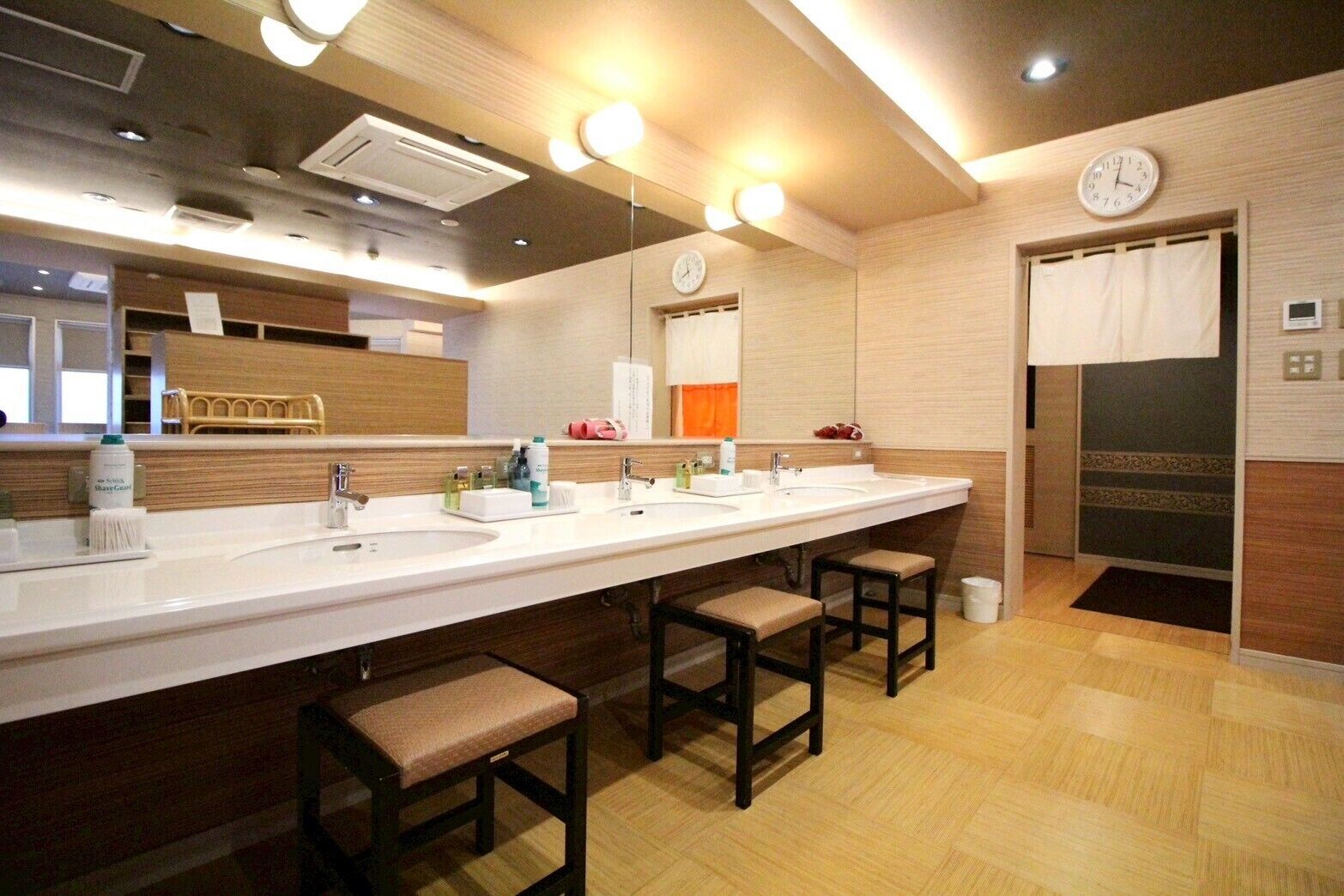A modern bathroom with a long vanity, multiple sinks, and a mirror. There are stools at the vanity and a clock on the wall.