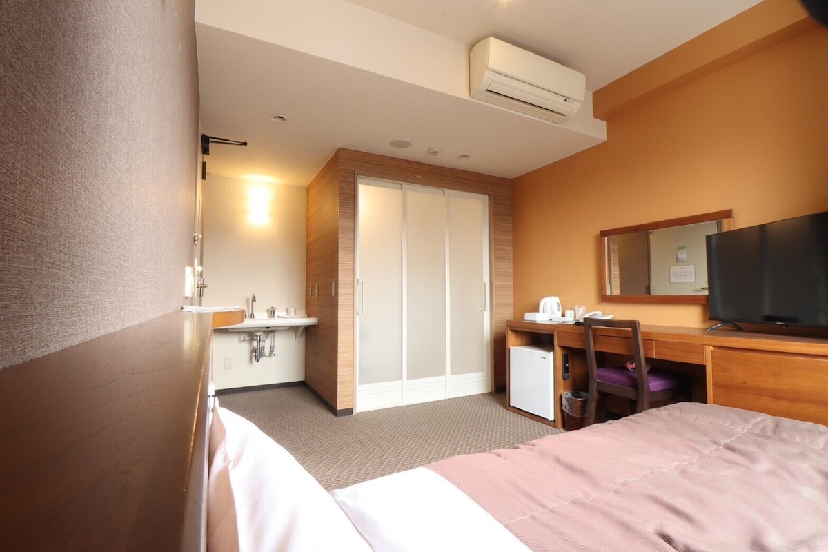 A hotel room with a bed, a desk with a chair, a television, a wardrobe, and a bathroom with a sink and mirror.