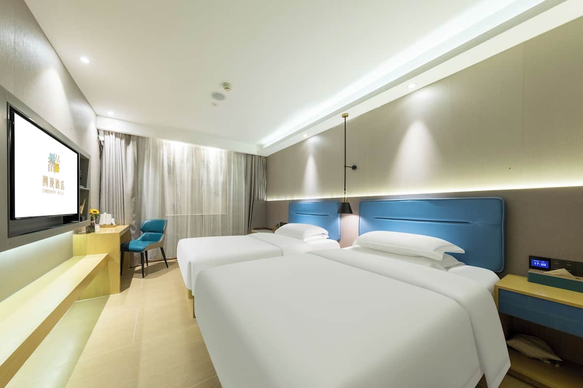 A hotel room with two beds, a desk, a chair, a TV, and a bedside table with a lamp.