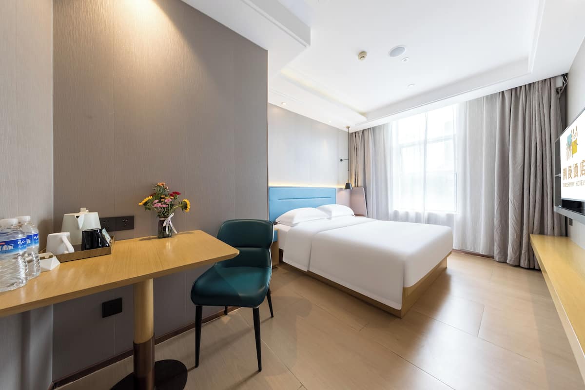 A hotel room with a bed, a desk with a chair, a TV, and a window with curtains.