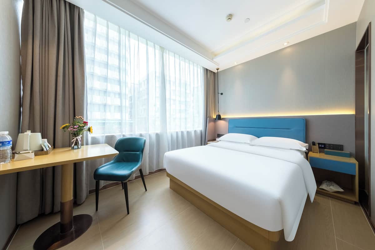 A hotel room with a bed, a desk, a chair, a nightstand, and a window with curtains.