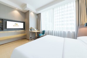 Ein Hotelzimmer mit einem großen Bett, einem Schreibtisch mit Stuhl, einem an der Wand montierten Fernseher und einem großen Fenster mit Vorhängen.