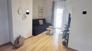 Olohuoneessa on sohva, pieni pyöreä pöytä, amppelissa roikkuva kasvi ja seinällä oleva kehystetty kasvitieteellinen taideteos.