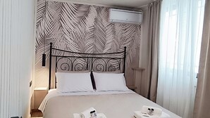 A neatly made bed with two pillows and two folded towels, a ceiling light fixture, and a wall-mounted air conditioner. - Skyline Penthouse, 5’ to Metro, Expansive Terrace,  Lightning-Fast Fiber WiFi (Athina)