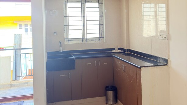 A kitchen with a black countertop, brown cabinets, a window, and a trash can. - SS Home Stay (Bengaluru)