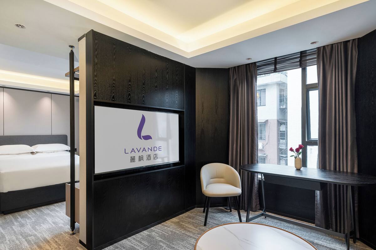 A modern hotel room with a bed, a desk, a chair, and a TV screen displaying the hotel's name.