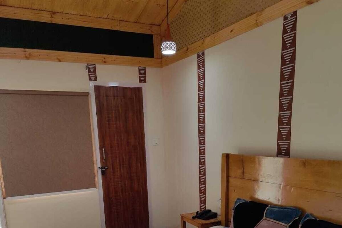 A bedroom with a wooden ceiling, a bed with a grey bedspread, a wooden headboard, a bedside table with a phone, and a door with a brown pattern.