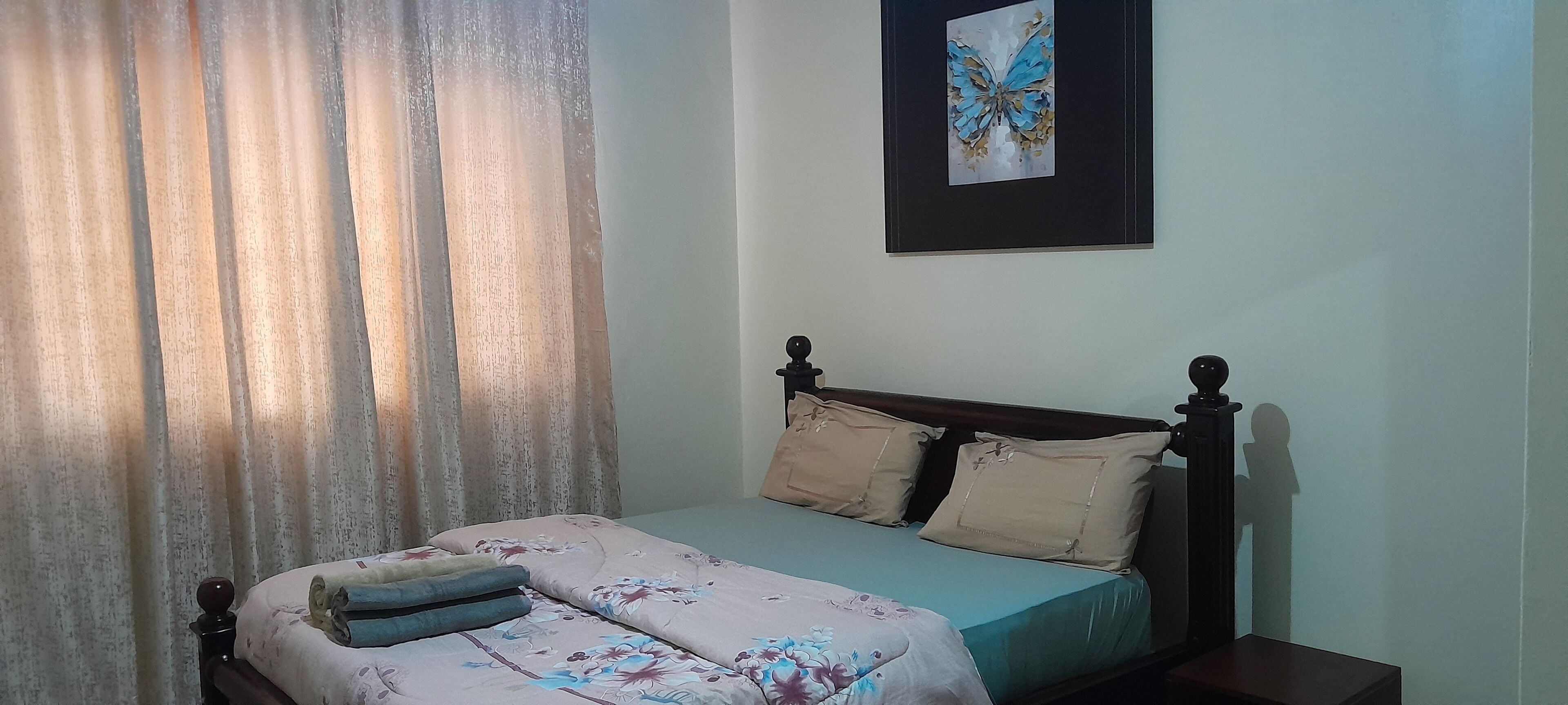 Une chambre à coucher avec un lit, des rideaux, un oreiller, un tableau décoratif et une table de chevet.