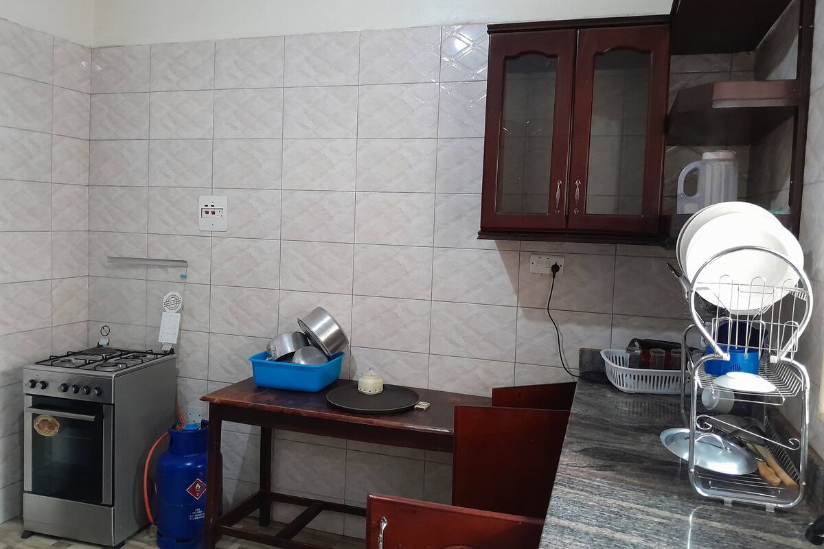 A kitchen with a gas stove, a blue gas cylinder, a wooden table, and a dish rack.