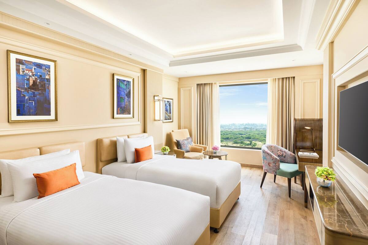 A hotel room with two beds, a desk, a TV, a chair, and a window with a city view.