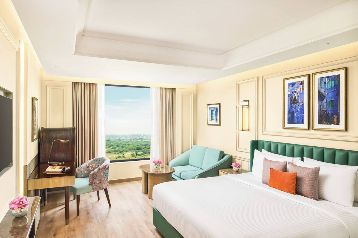 A hotel room with a bed, a desk, a chair, a small table, and a large window offering a view.