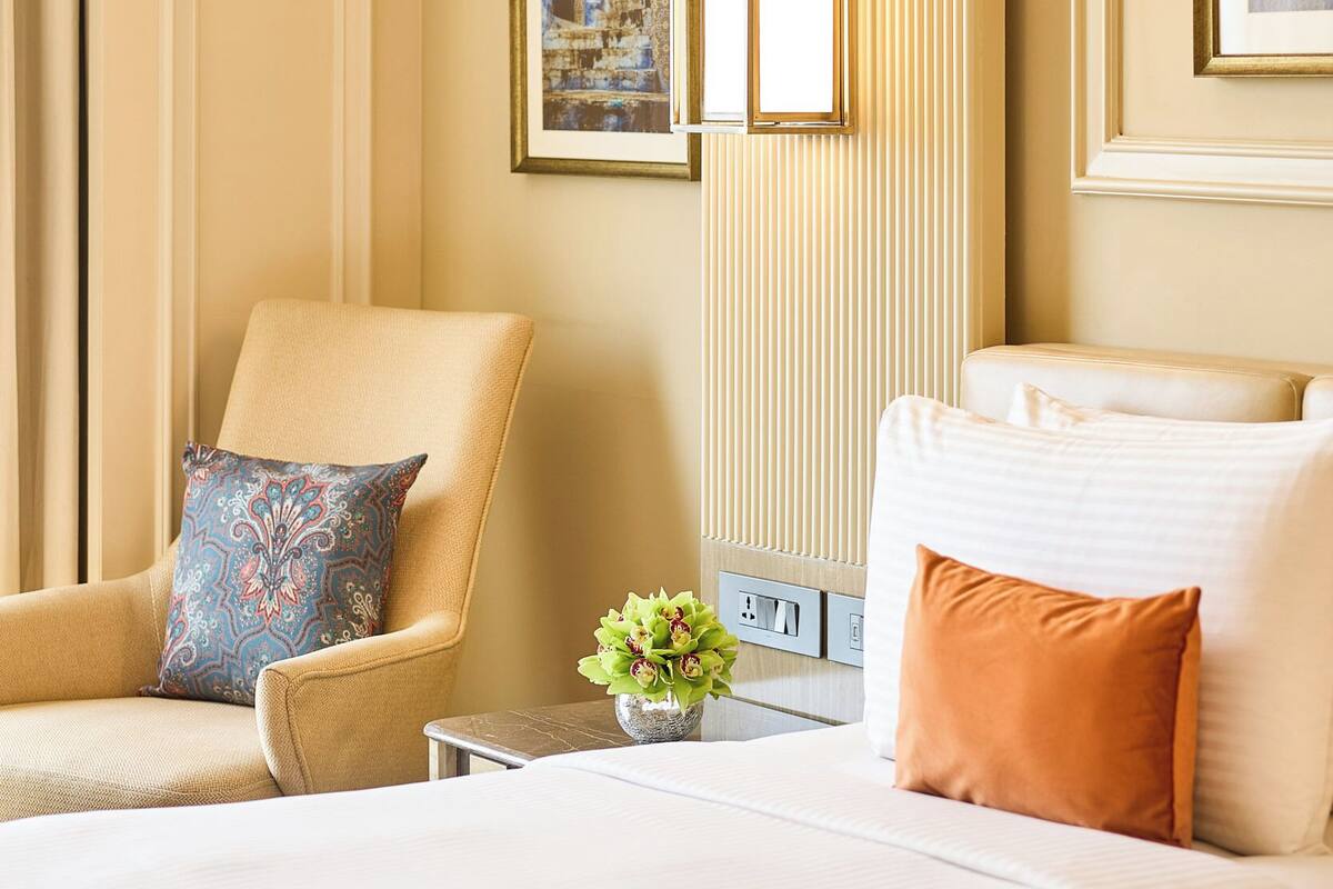 A hotel room with a bed, a chair, a table with a vase of flowers, and framed pictures on the walls.