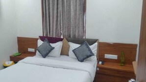 A neatly made bed with a wooden headboard, a bedside table with a green bottle, and a nightstand with a remote control. - HOTEL JASRAJ (Ahmedabad)