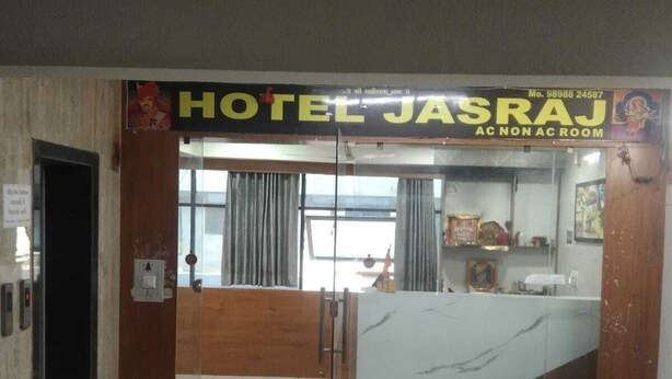 A hotel reception area with a marble counter, a reception desk, and a glass door leading to the lobby. - HOTEL JASRAJ (Ahmedabad)