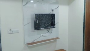 A room with a mounted flat-screen TV on a marble wall, a wooden shelf below it, and a door on the left. - HOTEL JASRAJ (Ahmedabad)