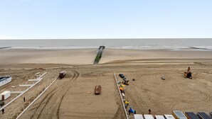 Een zandstrand met een grote groene constructie, mogelijk een windturbine, in het midden. Overal staan constructievoertuigen en materieel, en links een omheind gebied met gebouwen.