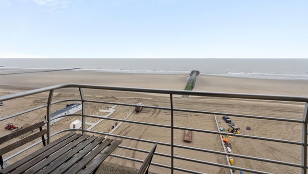 Uitzicht op een strand vanaf een balkon met een metalen hekwerk, een zandstrand en een heldere lucht.