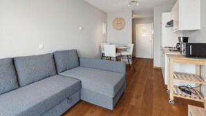 Een moderne woonkamer met een grijze hoekbank, een houten eettafel met stoelen, een keuken met een magnetron, en een klok aan de muur.