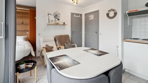 A compact room with a bed, a dining table with chairs, a kitchenette, and a small seating area with a chair and a blanket. - Nice studio near the beach (Blankenberge)