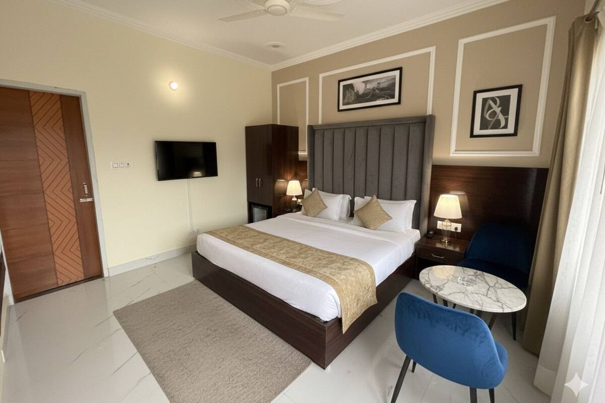 A hotel room with a large bed, a TV, a bedside table with a lamp, a small round table, and a blue chair.