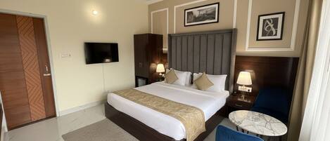 A hotel room with a large bed, a TV, a bedside table with a lamp, a small round table, and a blue chair.