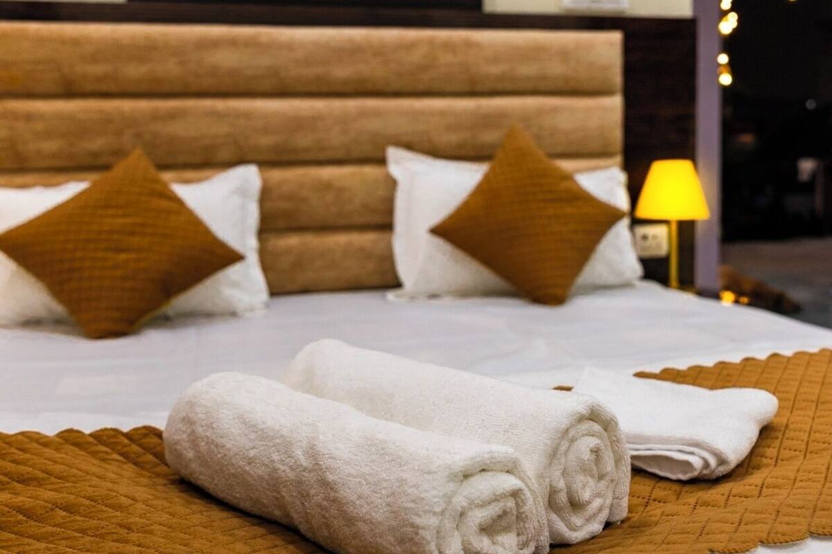 A neatly made bed with white linens and brown pillows, a painting on the wall, and a lamp on the side.