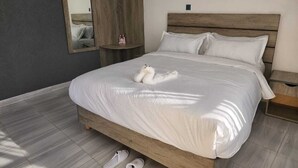 A neatly made bed with a white comforter and pillows, a wooden headboard, a wooden nightstand with a lamp, and a mirror on the wall. - Maara Lodge Chogoria (Chogoria)