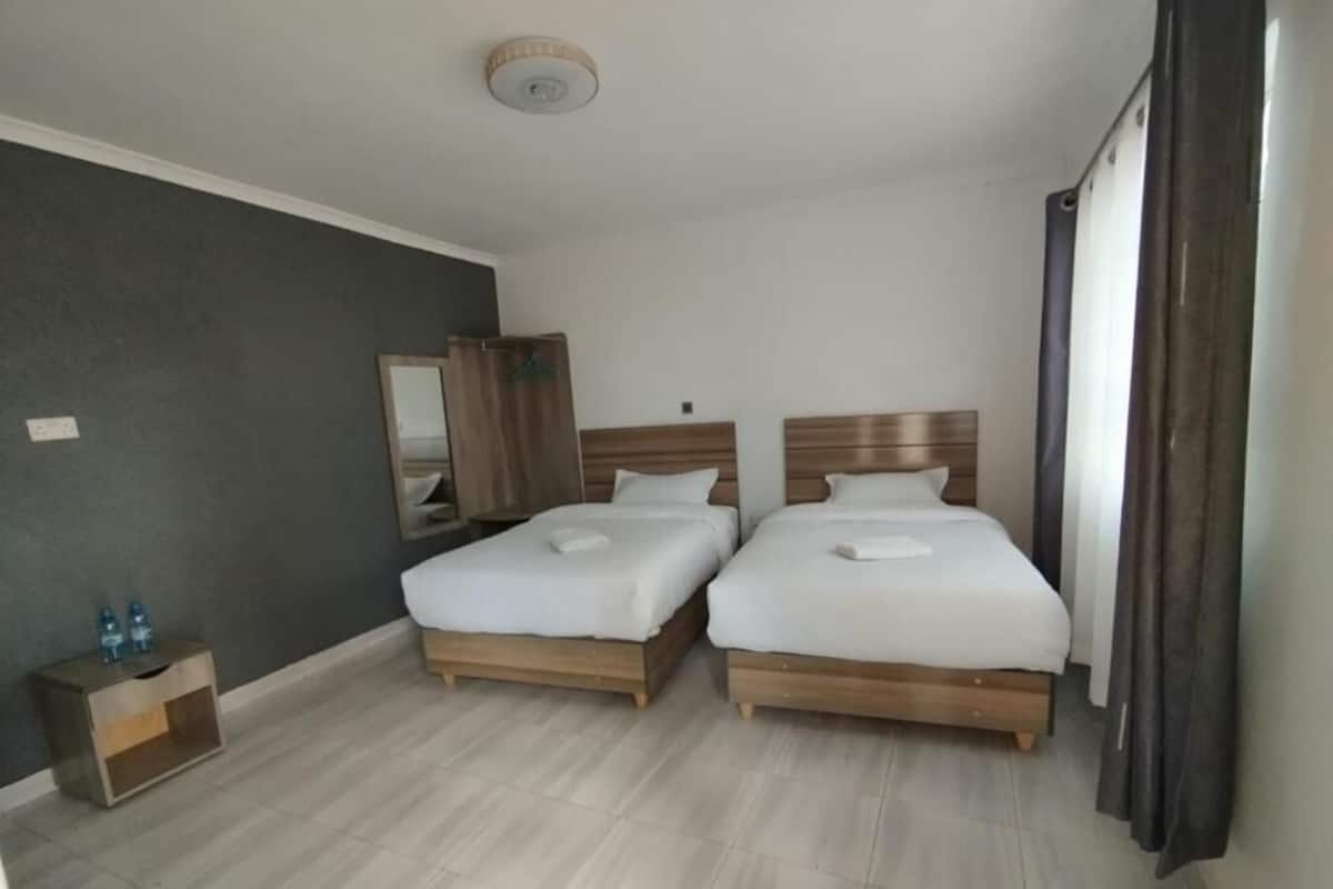 Habitación de hotel con dos camas, una mesita de noche con lámpara, un espejo y una ventana con cortinas.