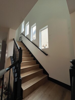 Una escalera con escalones de madera y barandillas negras, que lleva a un rellano con tres ventanas.