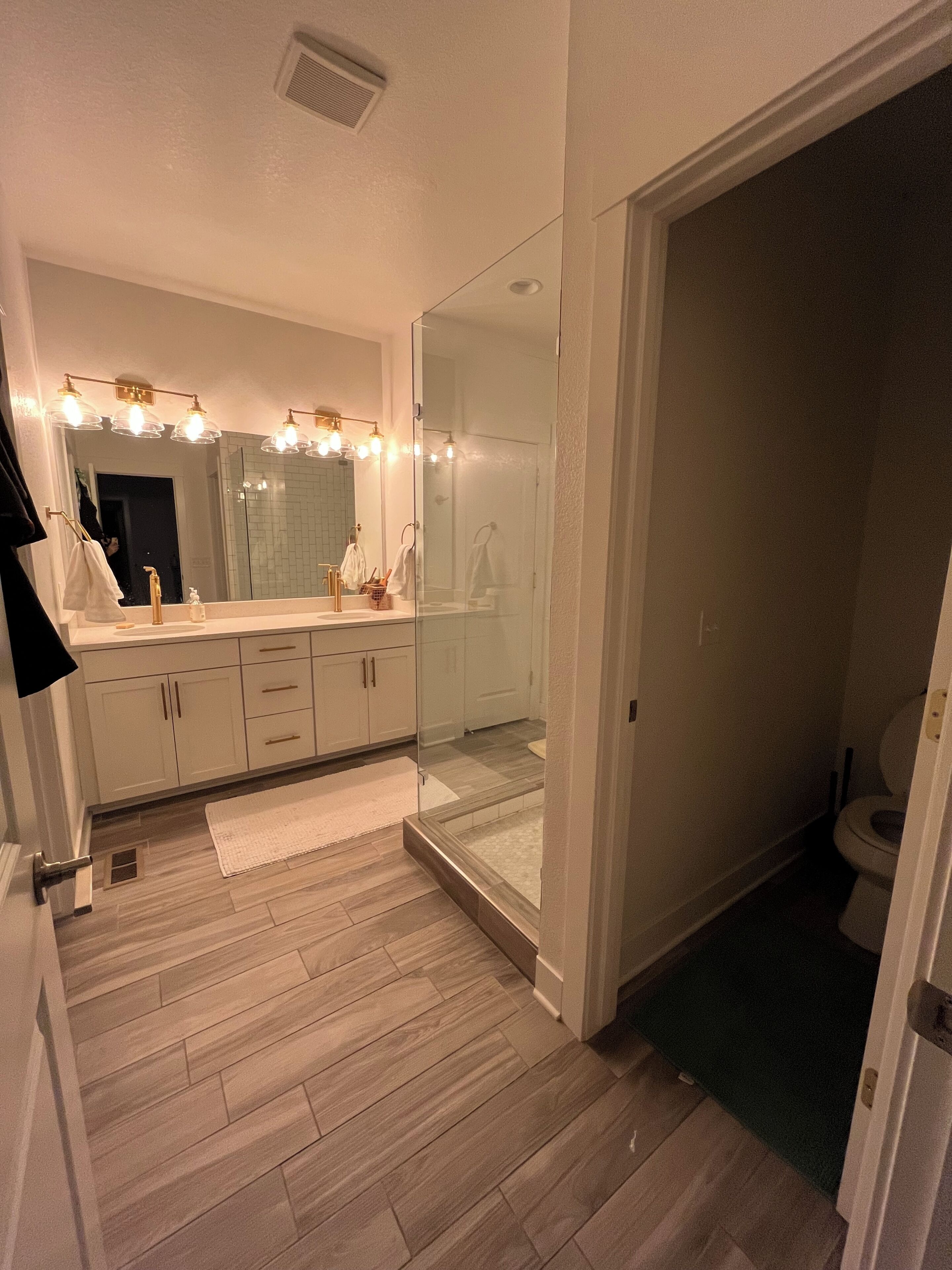 Un baño moderno con ducha envuelta en cristal, tocador con un espejo grande e iluminación superior, y suelo de madera.