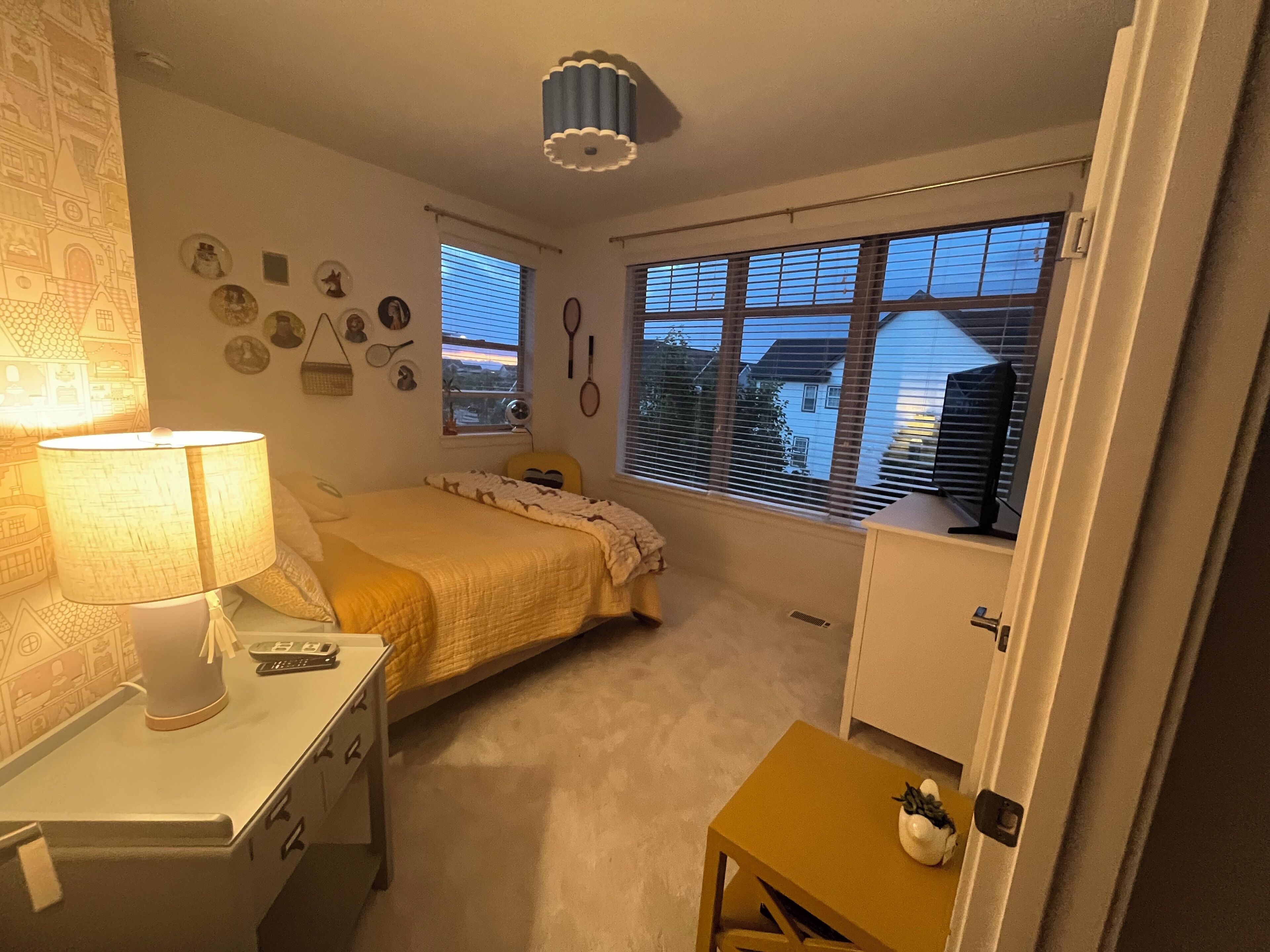 A bedroom with a bed, a desk with a lamp, a window with blinds, and a wall with various decorations.