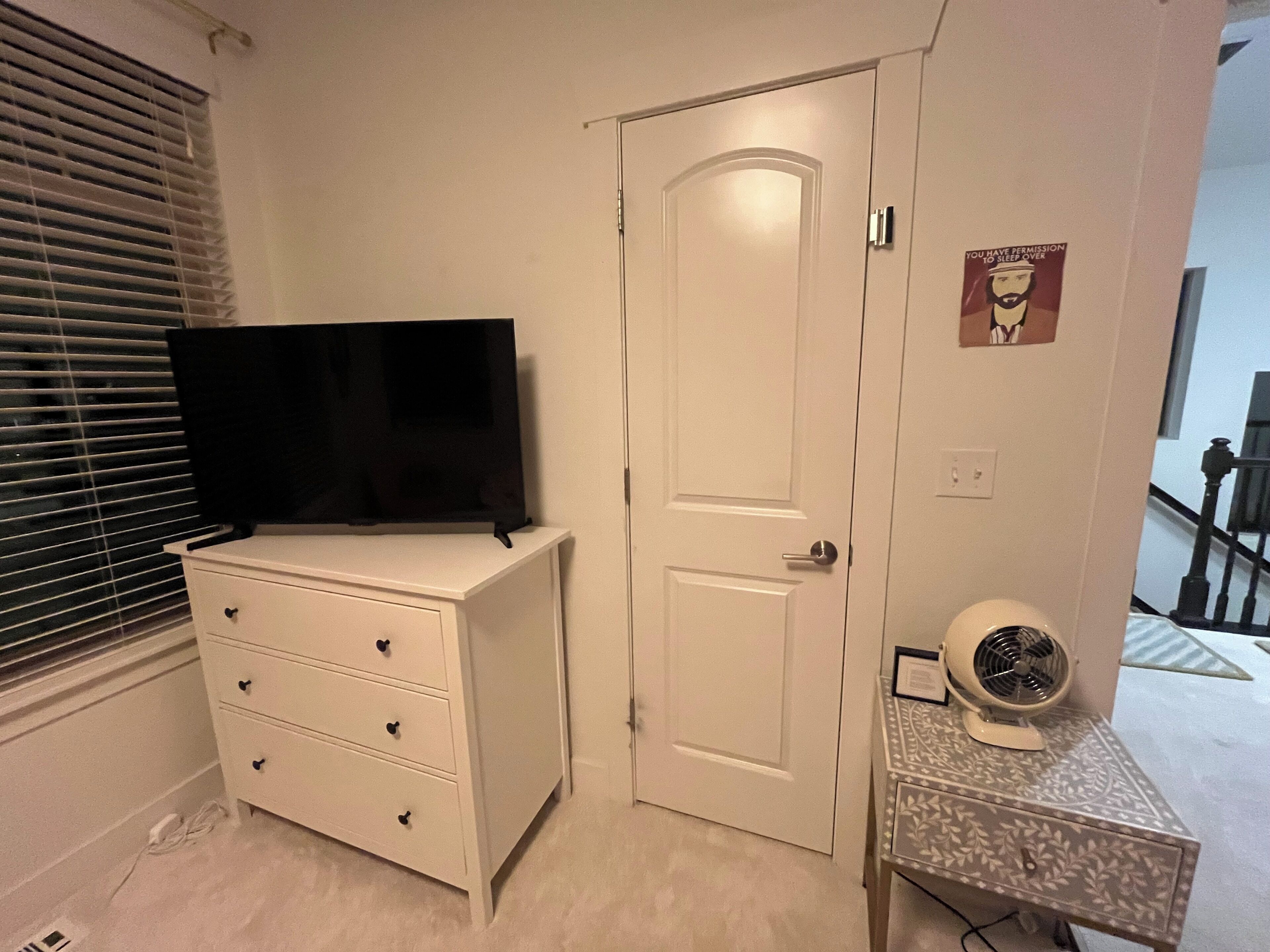A bedroom with a white dresser, a closed door, a wall-mounted fan, and a window with blinds.