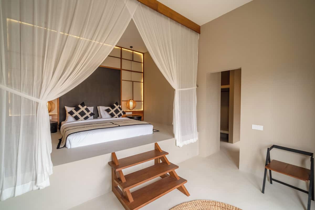 A bedroom with a bed, white curtains, a bedside table, a lamp, and a wooden bench.