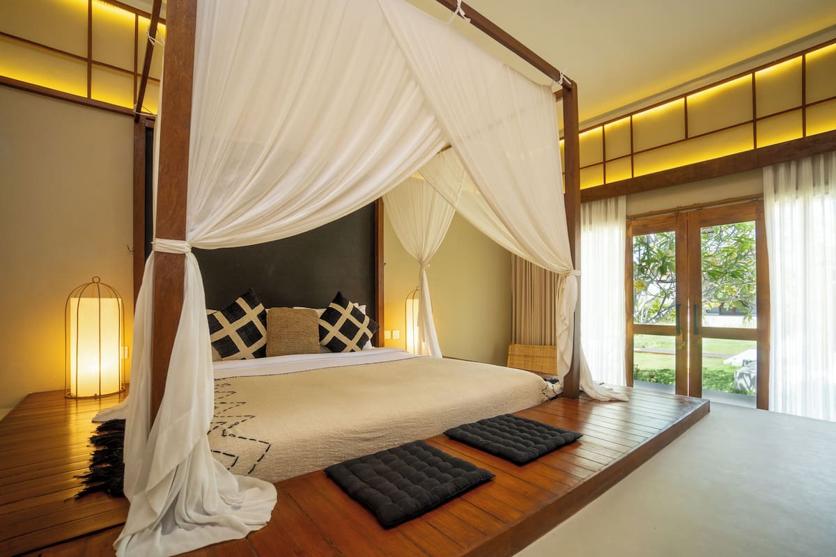 A spacious bedroom with a canopy bed, a view of greenery outside, and a wooden platform with two black cushions.