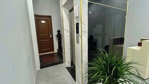 Un passadís amb paret de marbre, una porta de fusta amb la inscripció “001”, un telèfon i una planta en test.