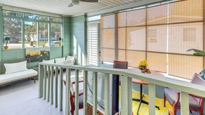 Una habitación con techo de madera, un sofá blanco, una mesa con sillas y ventanas grandes con persianas.