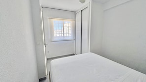 A small, narrow room with a single bed, a window with a grid pattern, and a door leading to another room.