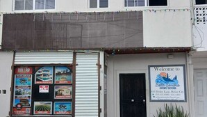 Un edificio de dos pisos con una persiana baja en la planta baja, un letrero que da la bienvenida a Bella Sandra Guest House y una pequeña maceta con plantas en la acera.