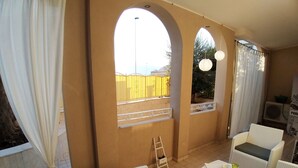 Un patio iluminado por el sol con mobiliario de mimbre blanco, una escalera y vista a una cerca amarilla a través de un ventanal arqueado.
