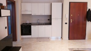 Una cocina pequeña con armarios blancos, un lavamanos y una estufa. Se ve una puerta de madera con un teléfono y un perchero en la pared.