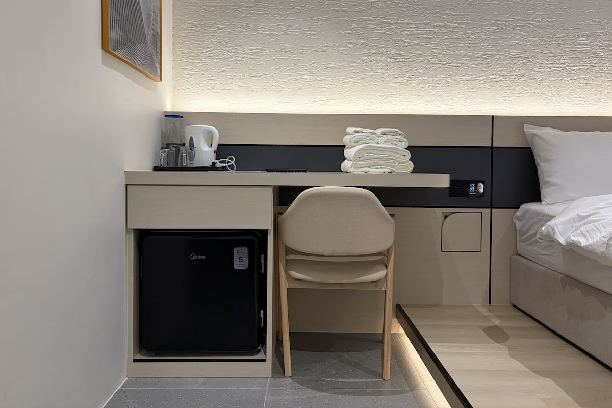 A compact hotel room with a bed, a desk with a computer, a small refrigerator, and a chair.