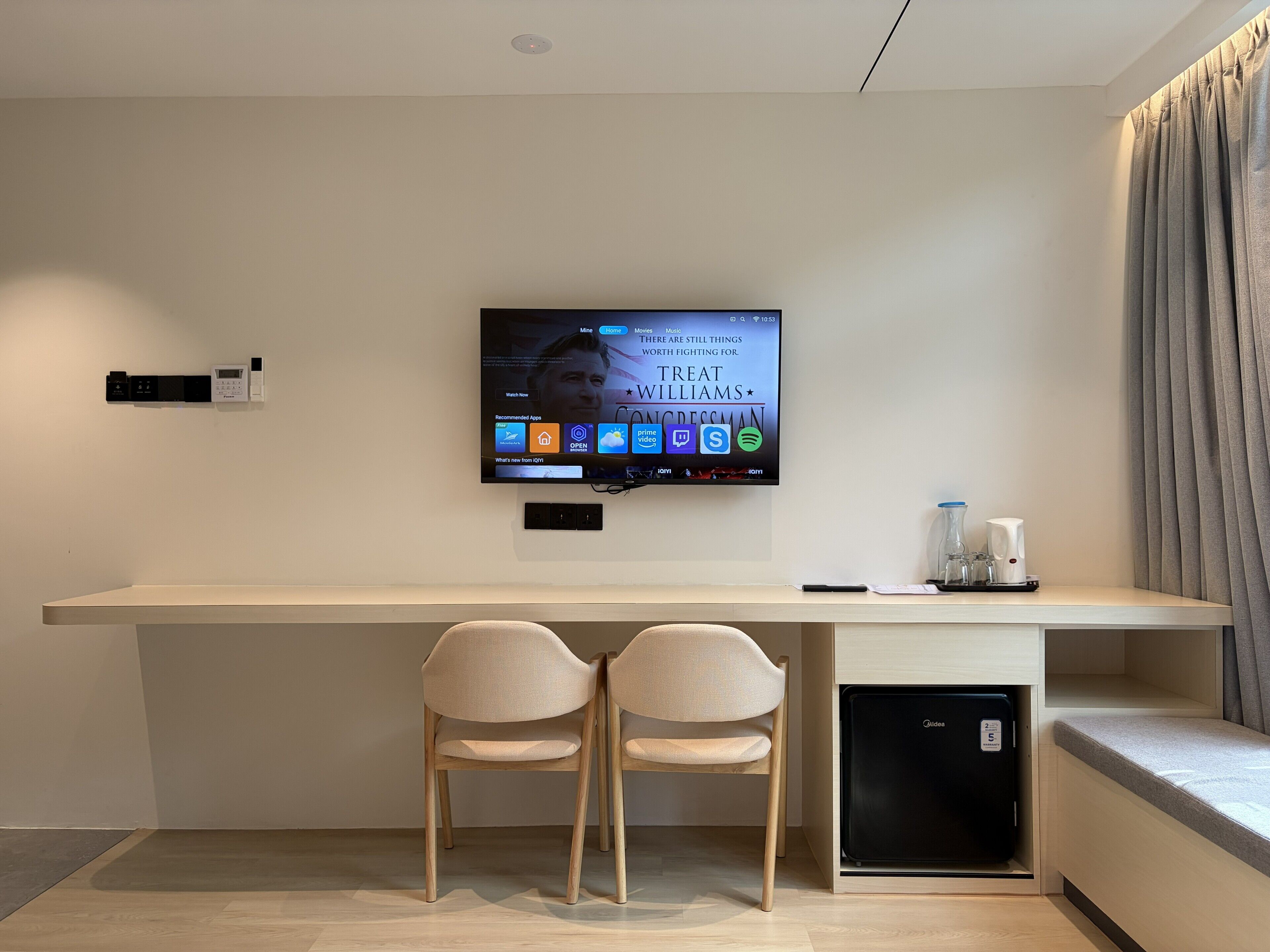 A modern hotel room with a long desk, two chairs, a flat-screen TV mounted on the wall, and a compact refrigerator.