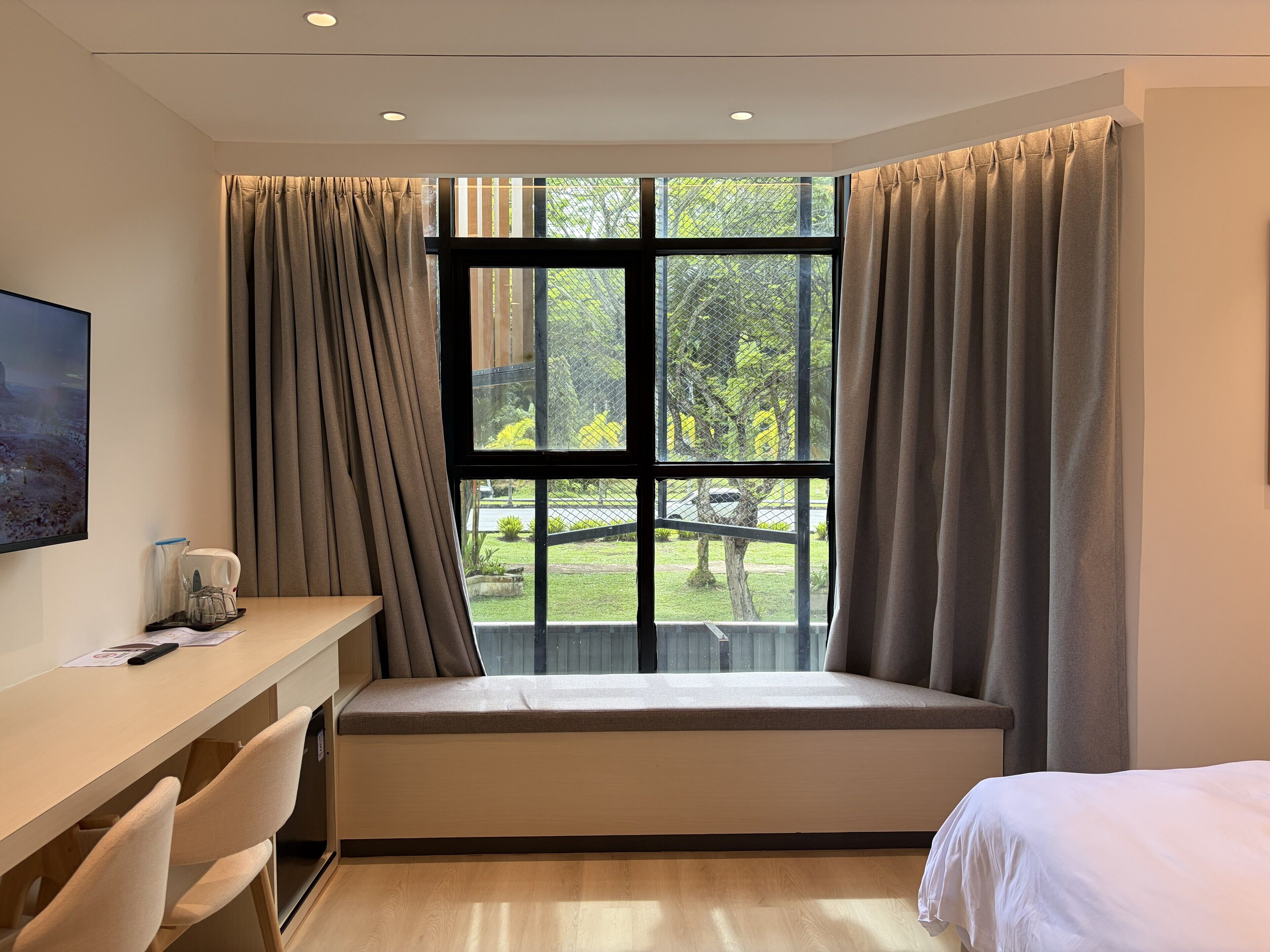 A hotel room with a large window, a desk with a chair, a bed, and a small table with a cup and papers.