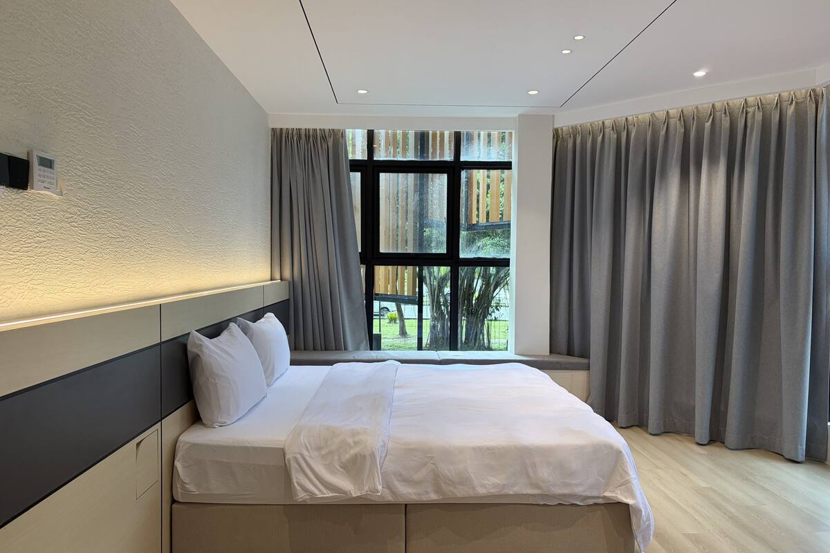 A modern bedroom with a large bed, a headboard, a window with curtains, and a view of greenery outside.