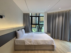 A modern bedroom with a large bed, a headboard, a window with curtains, and a view of greenery outside.