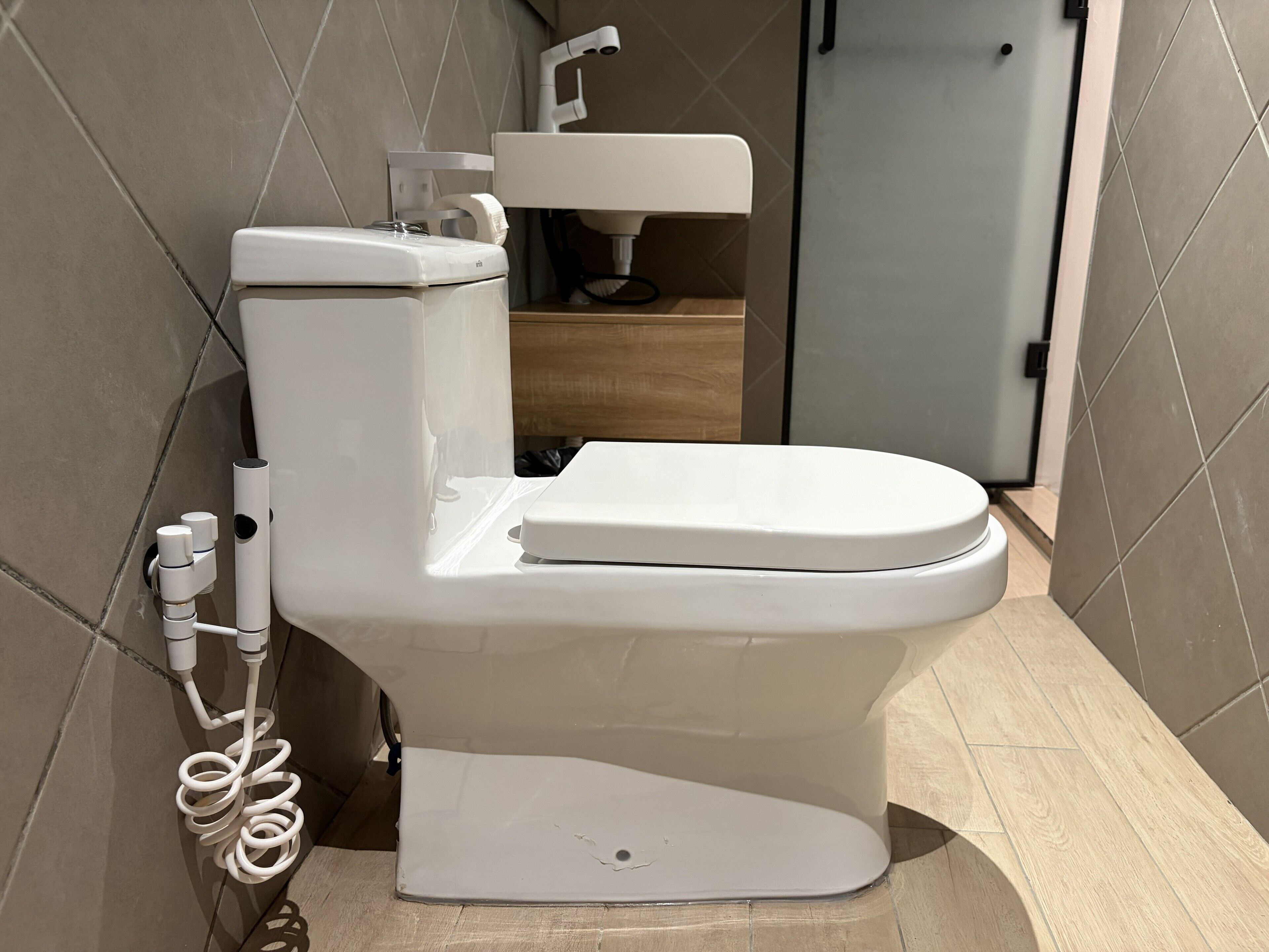 A modern toilet with a tank and seat, a coiled hose, and a wall-mounted faucet in a bathroom with tiled walls and a wooden floor.
